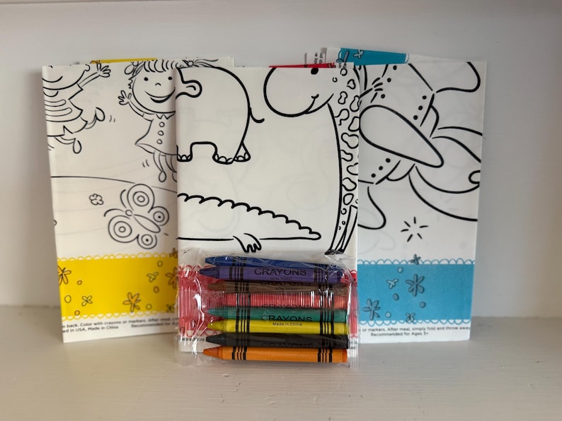 May include: Three coloring book pages with black line drawings of a girl, butterfly, elephant, giraffe, and flowers. A package of crayons in various colors sits in front of the pages. The pages have yellow and blue borders.