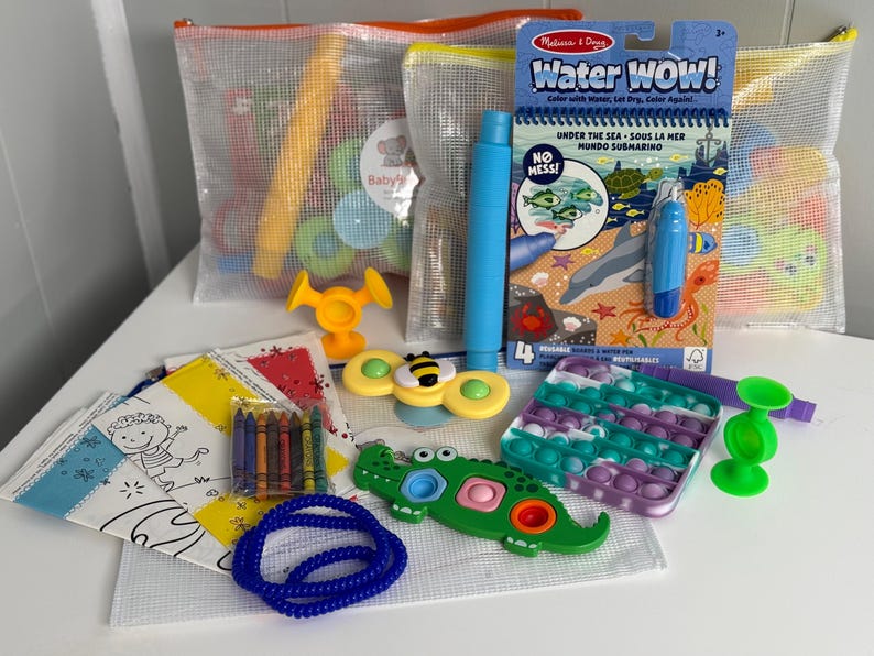 May include: A collection of children's toys, including a water-reveal coloring book, a pop-it, a fidget spinner, and crayons. The toys are in mesh bags and on a white surface. The coloring book says "Water WOW!" and "Under the Sea".