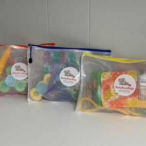 May include: Three mesh zipper bags filled with colorful toys. Each bag has a different colored zipper: orange, blue, and yellow. The bags contain various shapes and sizes of toys, including blocks and rings. Each bag has a round sticker with the text "BabyBusyBags".