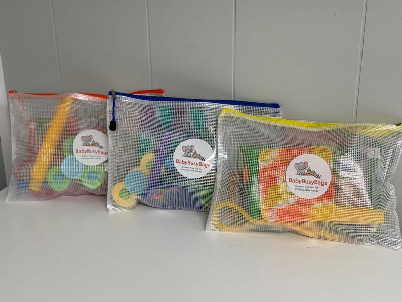 May include: Three mesh zipper bags filled with colorful toys. Each bag has a different colored zipper: orange, blue, and yellow. The bags contain various shapes and sizes of toys, including blocks and rings. Each bag has a round sticker with the text "BabyBusyBags".