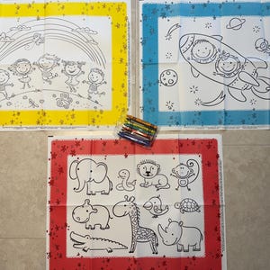 May include: Three coloring placemats with black outlines on white backgrounds. One features children under a rainbow, another a rocket ship with passengers, and the third various animals. A set of crayons is also visible.