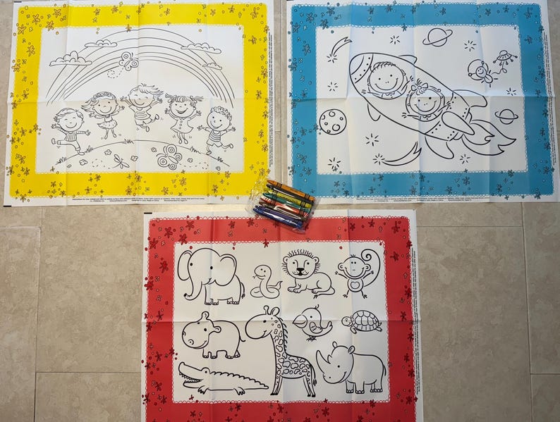 May include: Three coloring placemats with black outlines on white backgrounds. One features children under a rainbow, another a rocket ship with passengers, and the third various animals. A set of crayons is also visible.