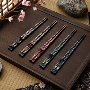 Peut inclure: Cinq paires de baguettes en bois avec une surface texturée et facettée. Chaque paire présente une couleur différente : noir, brun foncé, brun rougeâtre, vert foncé et bleu marine. Les manches sont ornés de petits motifs floraux dorés.