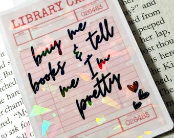 May include: A clear bookmark with a library card design. The text on the bookmark reads "buy me books & tell me I'm pretty" in black script. The bookmark has a holographic glitter effect and small heart graphics.