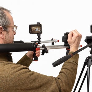May include: A person using a camera stabilizer rig with a smartphone and a camera mounted on a tripod. The rig has a black padded shoulder rest and red accents. The tripod is black.