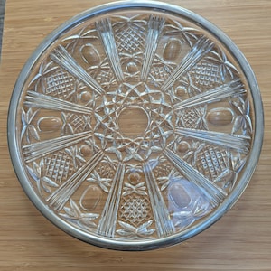 May include: A circular clear glass plate featuring a metallic silver rim and an intricate pressed pattern. The design includes radiating linear rays, diamond-shaped cross-hatching, and smooth oval indentations arranged in a symmetrical radial layout.