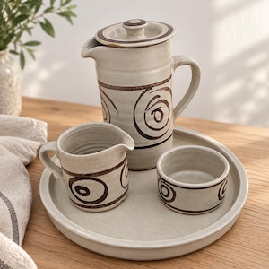 May include: A ceramic set featuring a lidded pitcher, a small creamer pitcher, and a small bowl, all arranged on a round tray. Each piece is off-white with dark brown hand-painted spiral patterns and a smooth, matte finish throughout the set.