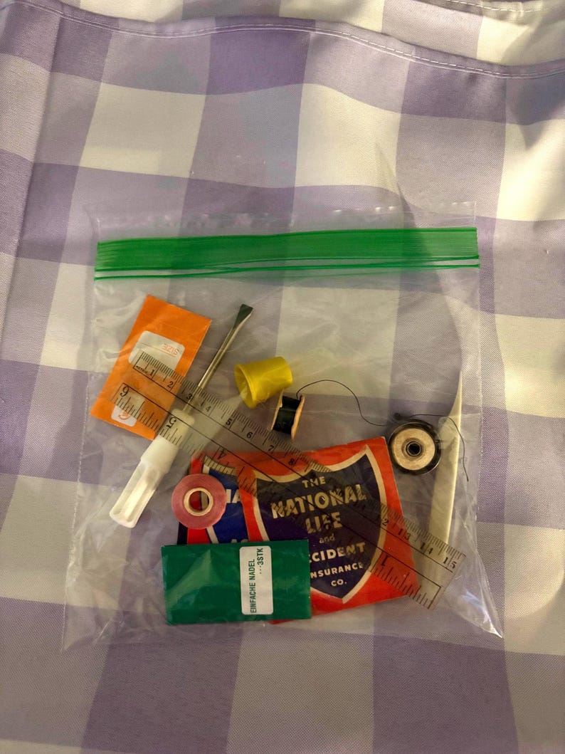 May include: A clear plastic bag containing sewing supplies including a small clear ruler with markings in inches, a yellow thimble, a black spool of thread, a metal seam ripper, a pair of tweezers, a pink button, and a vintage National Life insurance card.