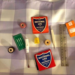 May include: A collection of sewing notions including two red matchbooks labeled The National Life and Accident Insurance Co., a green needle case, a yellow thimble, spools of thread, a white seam ripper, and a 6-inch metal ruler.