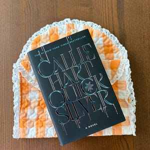 May include: A hardcover book titled "Quicksilver" by Callie Hart, with a black cover and silver lettering, rests on a quilted orange and white gingham tea cozy with a white lace trim. The book is a New York Times bestseller.