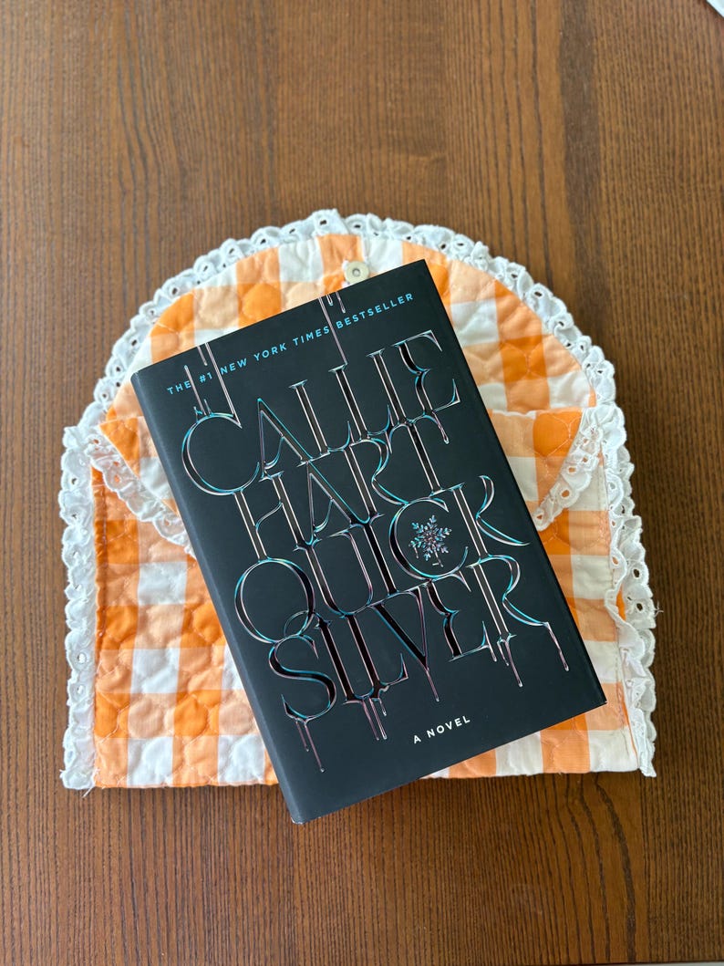 May include: A hardcover book titled "Quicksilver" by Callie Hart, with a black cover and silver lettering, rests on a quilted orange and white gingham tea cozy with a white lace trim. The book is a New York Times bestseller.