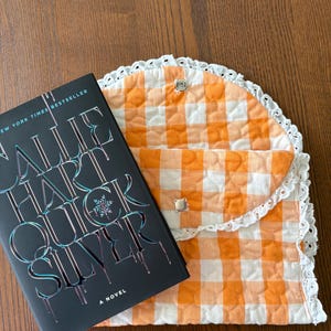 May include: A hardcover book titled "Quicksilver" by Sallie Hart next to an orange and white gingham quilted book sleeve with a white lace trim. The book sleeve has two silver snap closures.