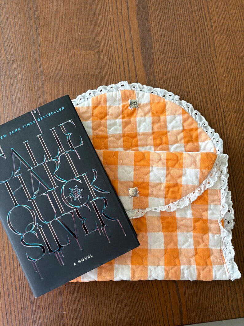 May include: A hardcover book titled "Quicksilver" by Sallie Hart next to an orange and white gingham quilted book sleeve with a white lace trim. The book sleeve has two silver snap closures.