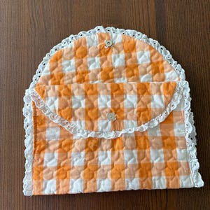 May include: A quilted, orange and white gingham clutch with a white lace trim. The clutch has a rounded flap that fastens with a small button. The fabric has a diamond pattern.