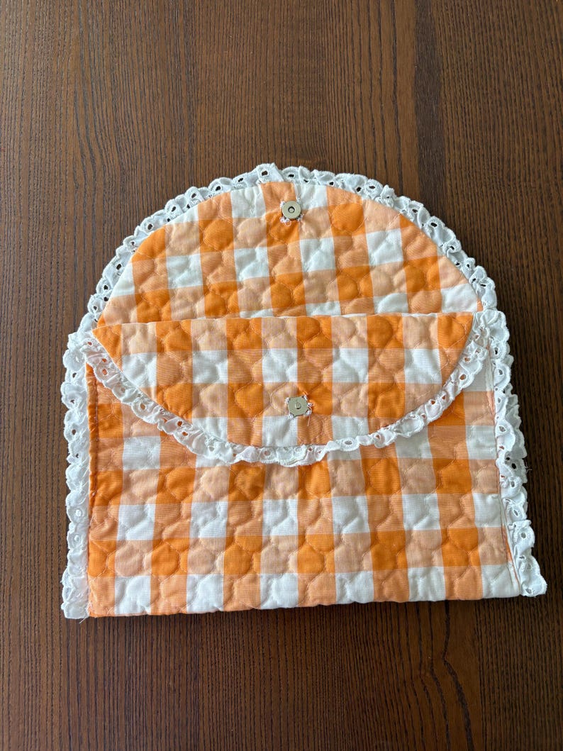 May include: A quilted, orange and white gingham clutch with a white lace trim. The clutch has a rounded flap that fastens with a small button. The fabric has a diamond pattern.