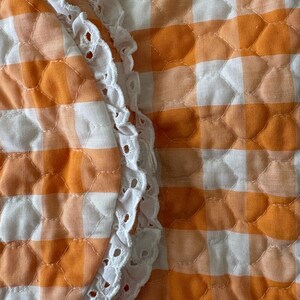 May include: Close-up of a quilted fabric with an orange and white gingham pattern. A white, scalloped lace trim borders the edge of the fabric, adding a delicate contrast to the bold pattern. The fabric has a textured, padded appearance.