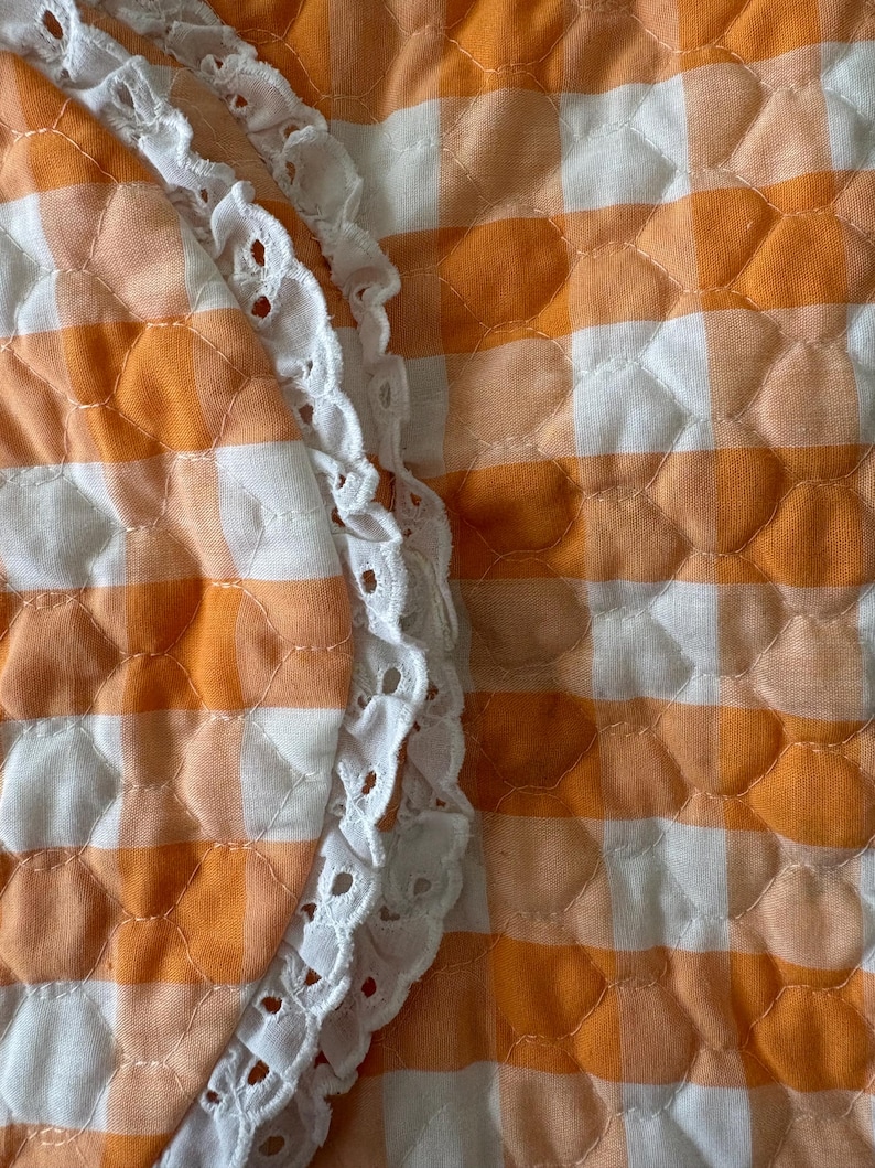 May include: Close-up of a quilted fabric with an orange and white gingham pattern. A white, scalloped lace trim borders the edge of the fabric, adding a delicate contrast to the bold pattern. The fabric has a textured, padded appearance.