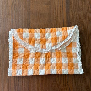 May include: A quilted envelope-style clutch in a vibrant orange and white gingham pattern. The edges are trimmed with delicate white lace, adding a touch of vintage charm. The clutch is closed with a flap.