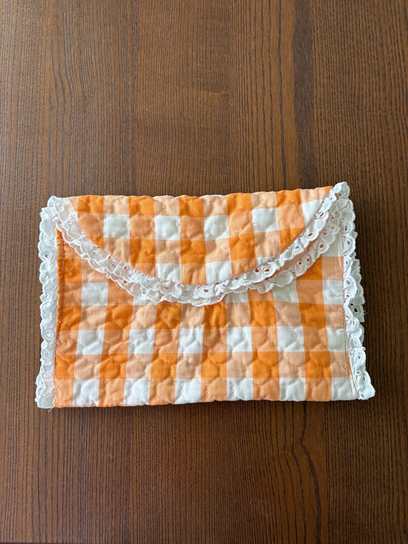 May include: A quilted envelope-style clutch in a vibrant orange and white gingham pattern. The edges are trimmed with delicate white lace, adding a touch of vintage charm. The clutch is closed with a flap.
