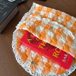 May include: An orange and white gingham quilted book sleeve with a white lace trim, holding a red book titled "A Court of Thorns and Roses". The sleeve is secured with two silver snap closures. Other books and e-readers are in the background.