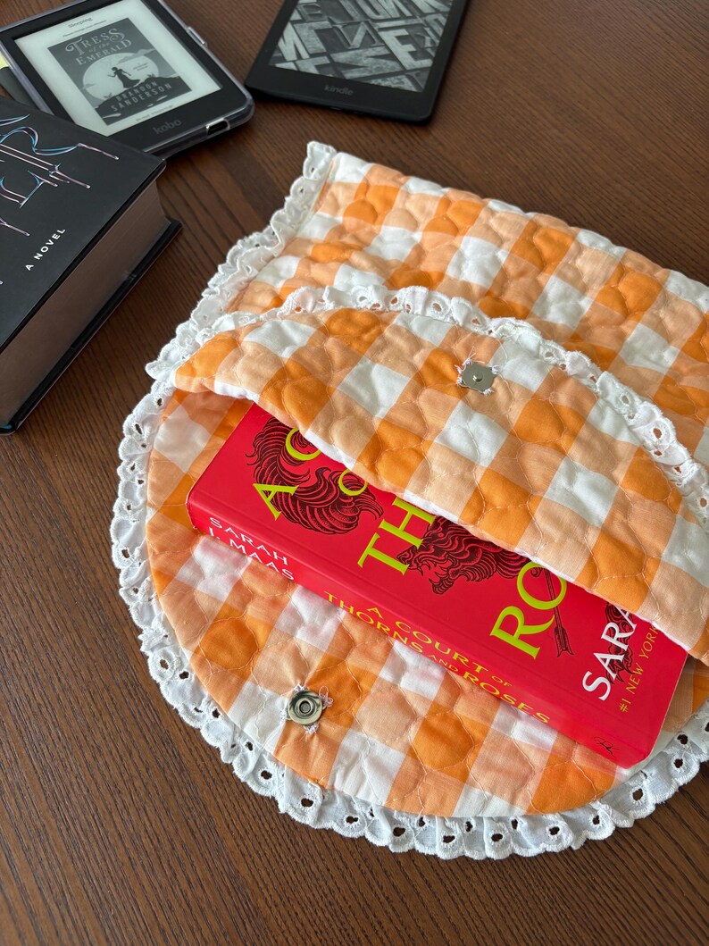 May include: An orange and white gingham quilted book sleeve with a white lace trim, holding a red book titled "A Court of Thorns and Roses". The sleeve is secured with two silver snap closures. Other books and e-readers are in the background.