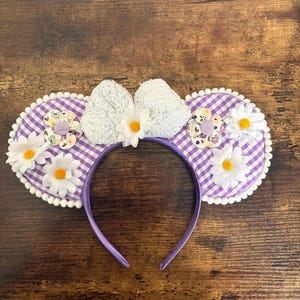 May include: A headband featuring two circular ears covered in a purple and white gingham fabric, trimmed with white pom-poms. A central white sequined bow is accented with several white daisy flowers with yellow centers and floral fabric circles.