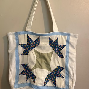 May include: A white fabric tote bag featuring a central quilt-style block pattern. The design includes four dark blue star shapes with small multi-colored floral prints, a central sage green square, and light blue fabric borders.