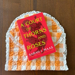 May include: A red book titled "A Court of Thorns and Roses" by Sarah J. Maas rests on an orange and white gingham quilted envelope. The envelope has a white lace trim. The book cover features a decorative illustration.
