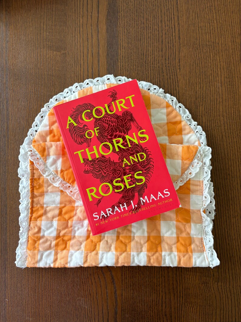 May include: A red book titled "A Court of Thorns and Roses" by Sarah J. Maas rests on an orange and white gingham quilted envelope. The envelope has a white lace trim. The book cover features a decorative illustration.