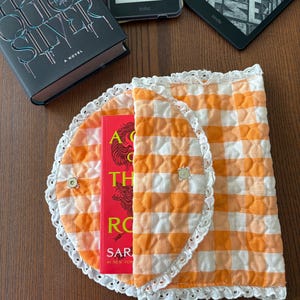 May include: An orange and white gingham book cover with a white lace trim, holding a red book. The cover is closed with two small buttons. A hardcover book and two e-readers are visible in the background.