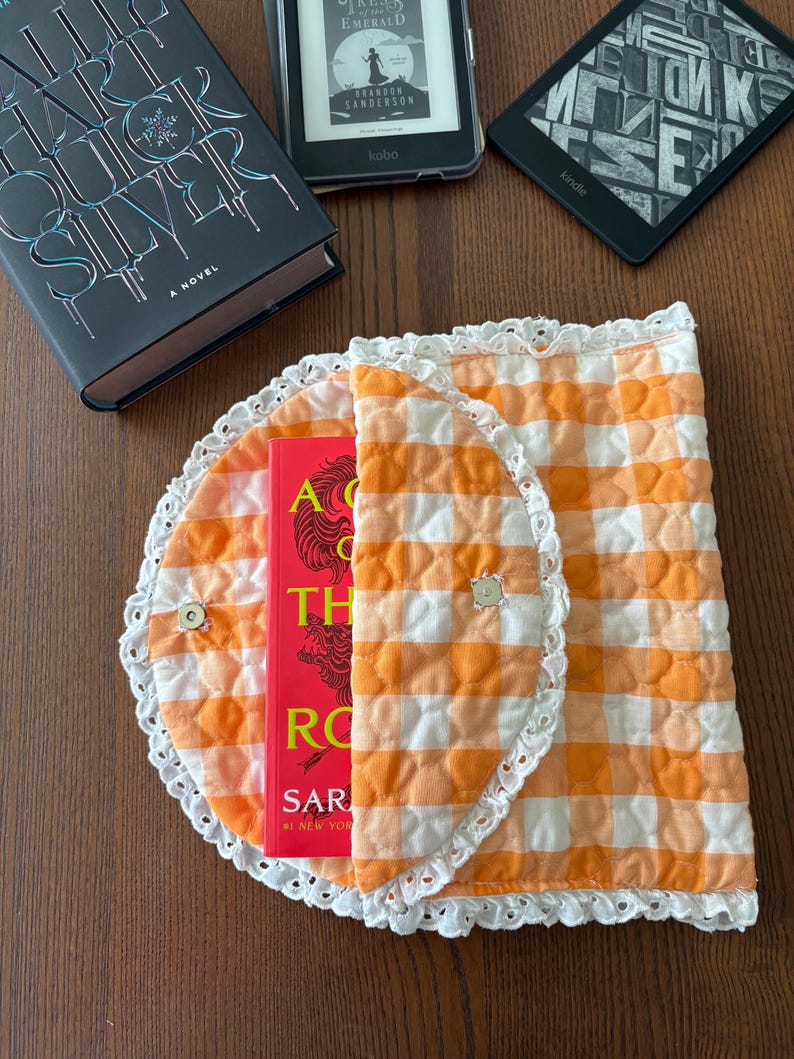 May include: An orange and white gingham book cover with a white lace trim, holding a red book. The cover is closed with two small buttons. A hardcover book and two e-readers are visible in the background.