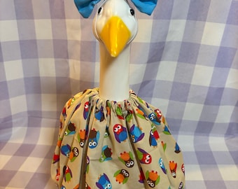 May include: A white goose figurine wearing a beige skirt with a colorful owl pattern and a blue fabric bow headband. The skirt features a gathered elastic waistband and a thin blue lace trim along the bottom edge, while the goose has a yellow beak.