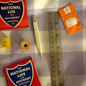 May include: A collection of sewing notions including two red and blue matchbook-style covers labeled The National Life and Accident Insurance Co., a yellow thimble, a wooden spool, a white seam ripper, and an orange needle packet next to a 6-inch ruler.