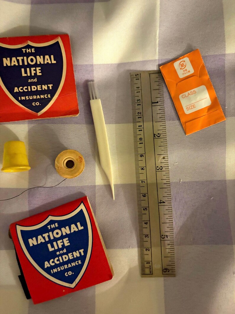 May include: A collection of sewing notions including two red and blue matchbook-style covers labeled The National Life and Accident Insurance Co., a yellow thimble, a wooden spool, a white seam ripper, and an orange needle packet next to a 6-inch ruler.