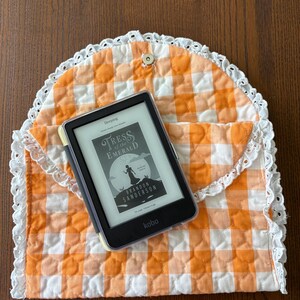 May include: An orange and white gingham quilted e-reader case with a white lace trim. The case holds a black e-reader displaying the book cover "Tress of the Emerald Sea" by Brandon Sanderson. The case is closed with a snap.