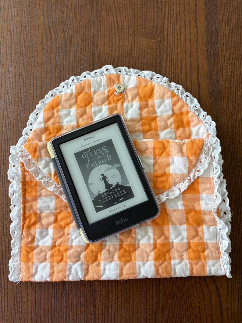 May include: An orange and white gingham quilted e-reader case with a white lace trim. The case holds a black e-reader displaying the book cover "Tress of the Emerald Sea" by Brandon Sanderson. The case is closed with a snap.