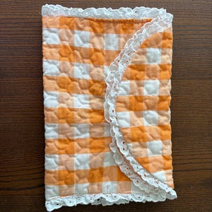 May include: A quilted, rectangular item with a gingham pattern in orange and white. The edges are trimmed with white lace. The item is folded in half, with the lace trim visible on the outer edge.