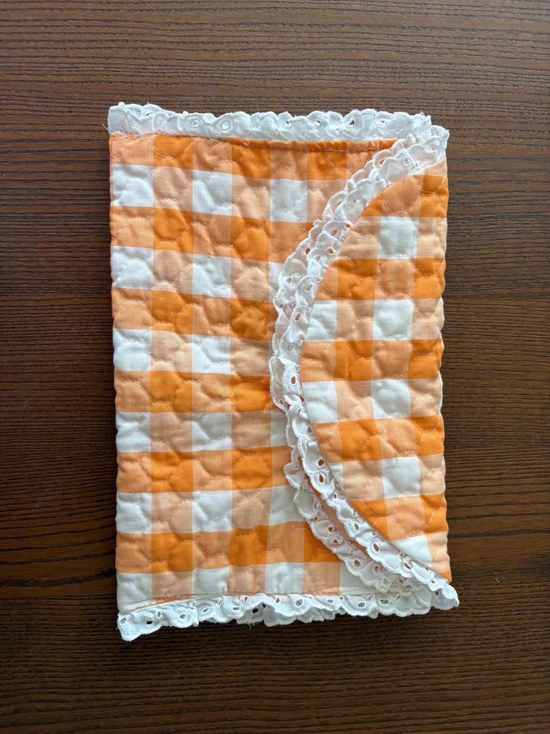 May include: A quilted, rectangular item with a gingham pattern in orange and white. The edges are trimmed with white lace. The item is folded in half, with the lace trim visible on the outer edge.