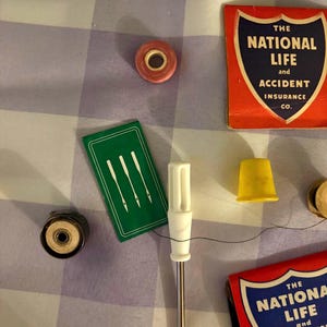 May include: A collection of sewing notions including a small pink spool, a green needle case with three white needle illustrations, a white-handled screwdriver, a yellow thimble, and two red matchbooks labeled The National Life and Accident Insurance Co.