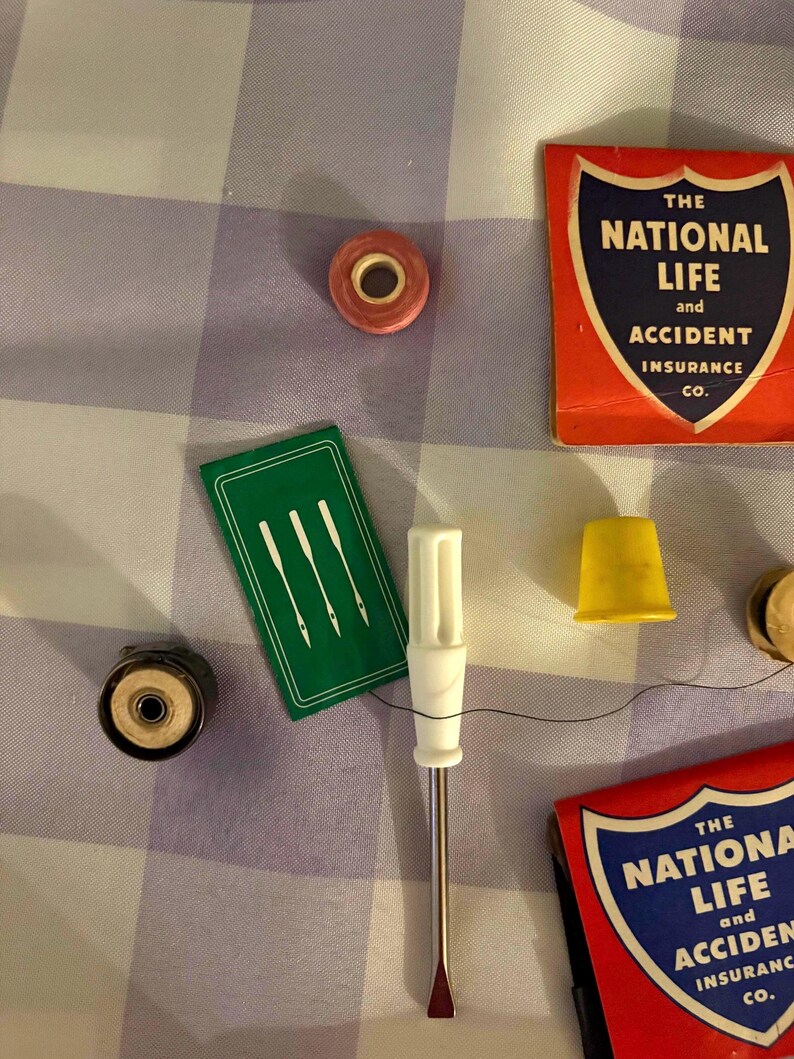 May include: A collection of sewing notions including a small pink spool, a green needle case with three white needle illustrations, a white-handled screwdriver, a yellow thimble, and two red matchbooks labeled The National Life and Accident Insurance Co.