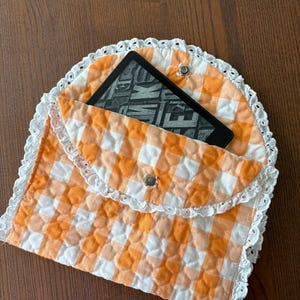 May include: An orange and white gingham quilted e-reader case with a white lace trim. The case is open, revealing a black e-reader with the word "WAKE" visible on the screen. The case is secured with a silver snap closure.