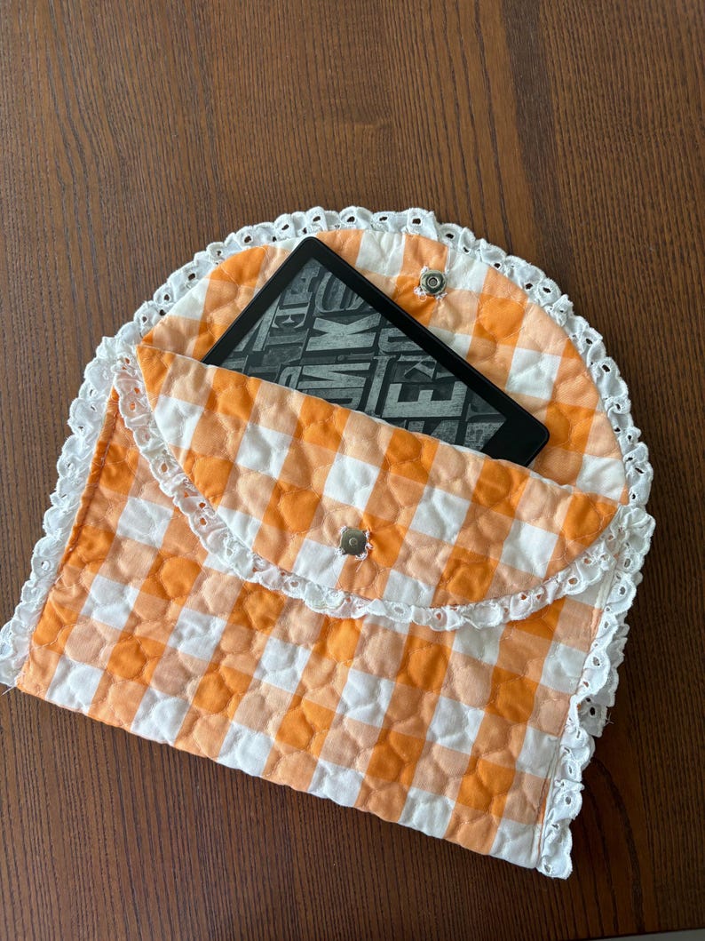 May include: An orange and white gingham quilted e-reader case with a white lace trim. The case is open, revealing a black e-reader with the word "WAKE" visible on the screen. The case is secured with a silver snap closure.