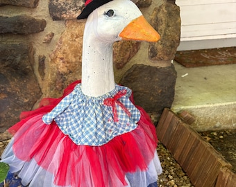 May include: A white goose lawn ornament wearing a black hat with a red band, a blue and white checkered collar with a small red bow, and a layered tutu skirt featuring alternating panels of red, white, and blue tulle fabric.