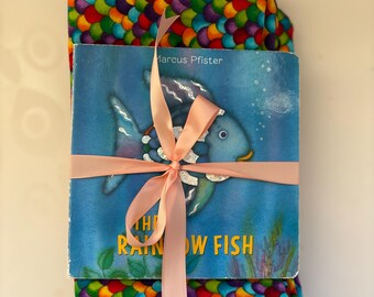 May include: A children's book titled "The Rainbow Fish" is wrapped with a pink ribbon and placed on top of a pair of leggings. The leggings feature a colorful scale pattern in shades of red, orange, yellow, green, blue, and purple.