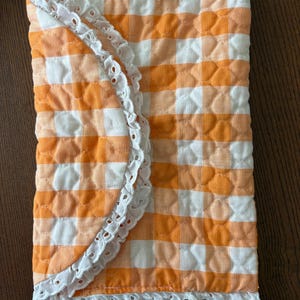 May include: A quilted, rectangular item with an orange and white gingham pattern. The edges are trimmed with a white, scalloped lace. The item appears to be a baby blanket or a similar textile product.