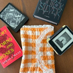 May include: An orange and white gingham fabric wrap with white lace trim, surrounded by books and e-readers. The books include "Callie Hart Quick Silver" and "A Court of Thorns and Roses" by Sarah J. Maas. Two e-readers are also visible.