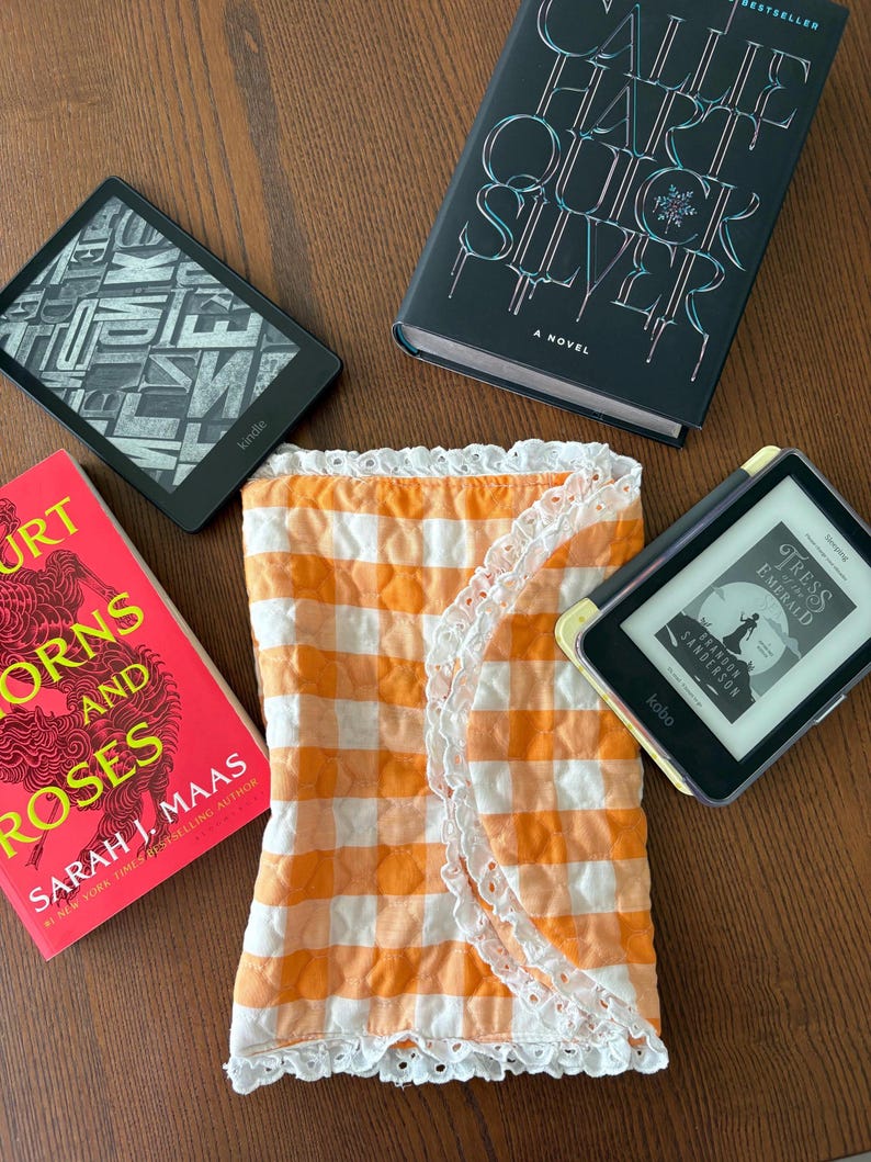 May include: An orange and white gingham fabric wrap with white lace trim, surrounded by books and e-readers. The books include "Callie Hart Quick Silver" and "A Court of Thorns and Roses" by Sarah J. Maas. Two e-readers are also visible.