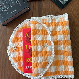 May include: An orange and white gingham book sleeve with a white lace trim, holding a red book. The sleeve is open, revealing the book's title. E-readers and a hardcover book are also visible.