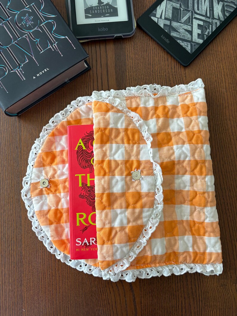 May include: An orange and white gingham book sleeve with a white lace trim, holding a red book. The sleeve is open, revealing the book's title. E-readers and a hardcover book are also visible.