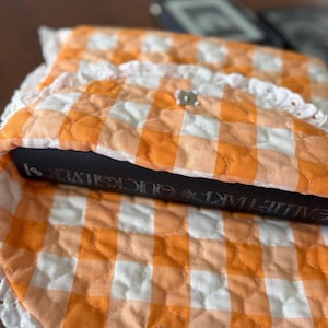 May include: An orange and white gingham quilted book sleeve with a white lace trim. The sleeve is closed with a silver snap. A book with the title "GALILEO'S TART QUICKSILVER" is inside the sleeve.